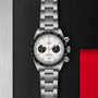 A TUDOR Black Bay Chrono with a stainless steel case, white dial, black subdials, and metal bracelet is showcased on a textured black and red surface.