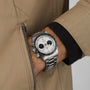 A person wearing a TUDOR Black Bay Chrono wristwatch with a white dial and black subdials, partially hidden by the cuff of a tan jacket.