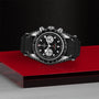The TUDOR Black Bay Chrono wristwatch, featuring a black strap and a black and silver design, is displayed on a glossy red and gray surface against a textured black background.
