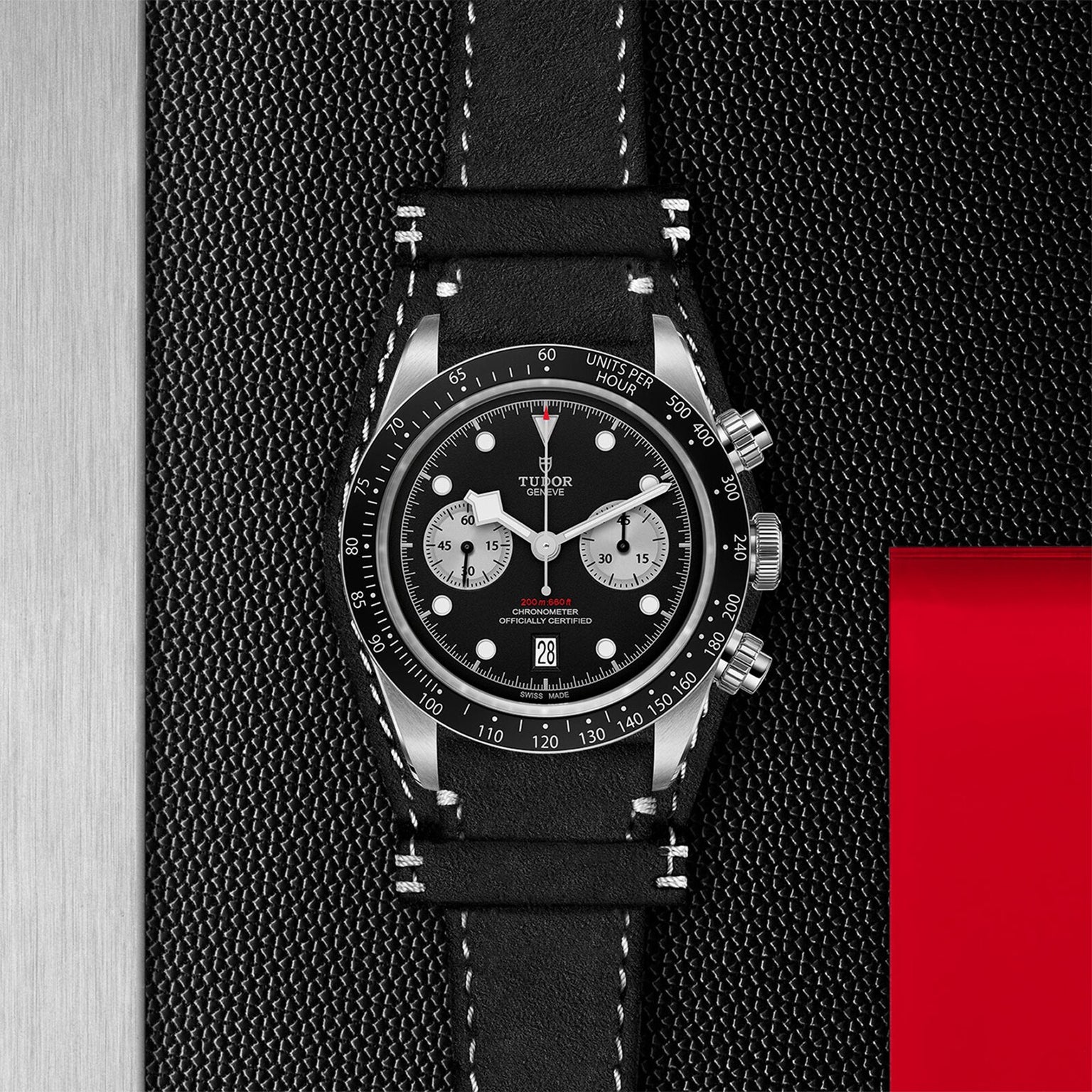 A TUDOR Black Bay Chrono chronograph wristwatch with a leather strap is shown on a textured black surface next to a red rectangular accent.