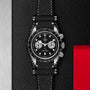 A TUDOR Black Bay Chrono chronograph wristwatch with a leather strap is shown on a textured black surface next to a red rectangular accent.