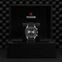 A TUDOR Black Bay Chrono wristwatch with a black and silver dial is showcased in an open black box bearing the TUDOR logo and shield emblem inside the lid.