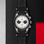 A TUDOR Black Bay Chrono wristwatch with a tachymetric scale, black leather strap, and white dial rests on a textured black surface featuring a metallic strip and a red rectangular accent.