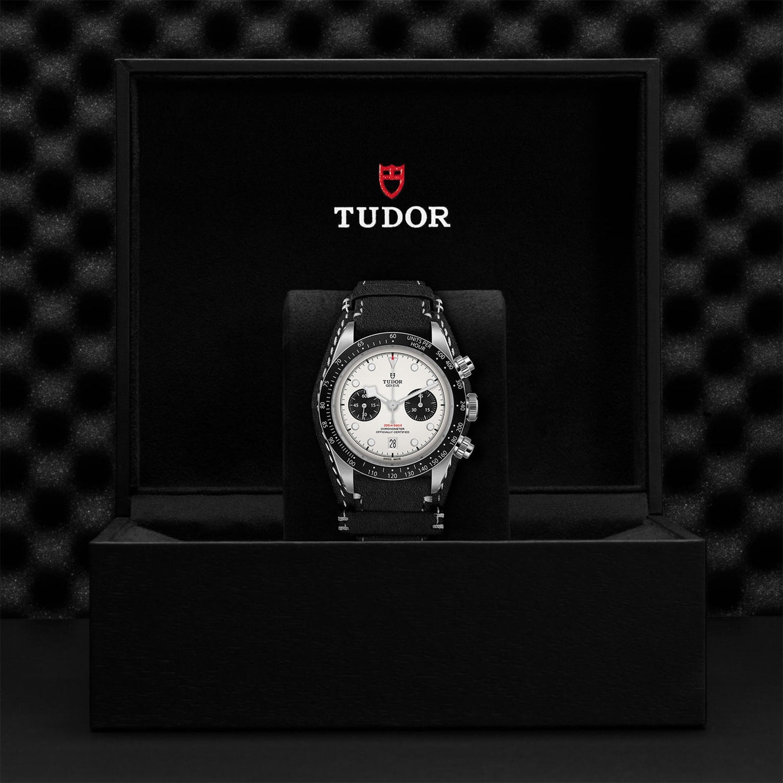 A TUDOR Black Bay Chrono wristwatch with a black strap and white dial is shown in an open black TUDOR box against a textured black backdrop, highlighting its signature tachymetric scale.