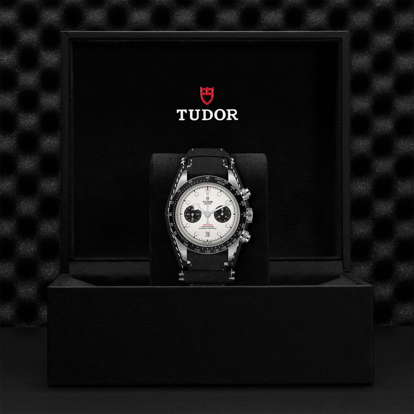 A TUDOR Black Bay Chrono wristwatch with a black strap and white dial is shown in an open black TUDOR box against a textured black backdrop, highlighting its signature tachymetric scale.