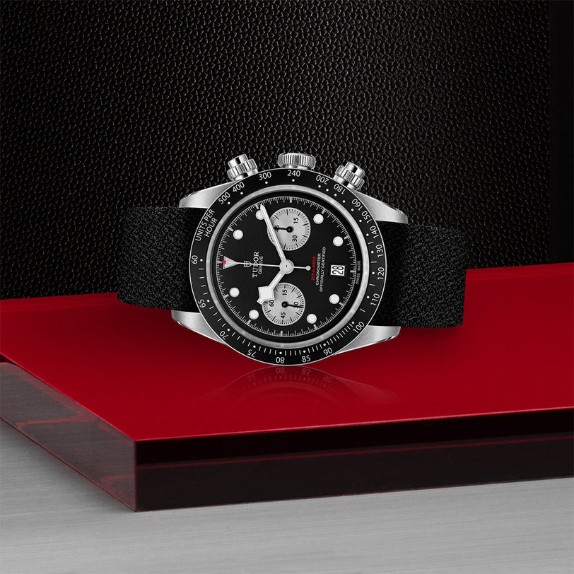 The TUDOR Black Bay Chrono, a black and silver chronograph wristwatch with a black strap, is displayed on a red glass surface against a textured black background.