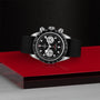 The TUDOR Black Bay Chrono, a black and silver chronograph wristwatch with a black strap, is displayed on a red glass surface against a textured black background.
