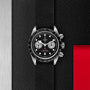 The TUDOR Black Bay Chrono features a black bezel, black textured strap, and two silver subdials, set against a black textured background with bold red and silver accents—a striking chronograph from TUDOR.