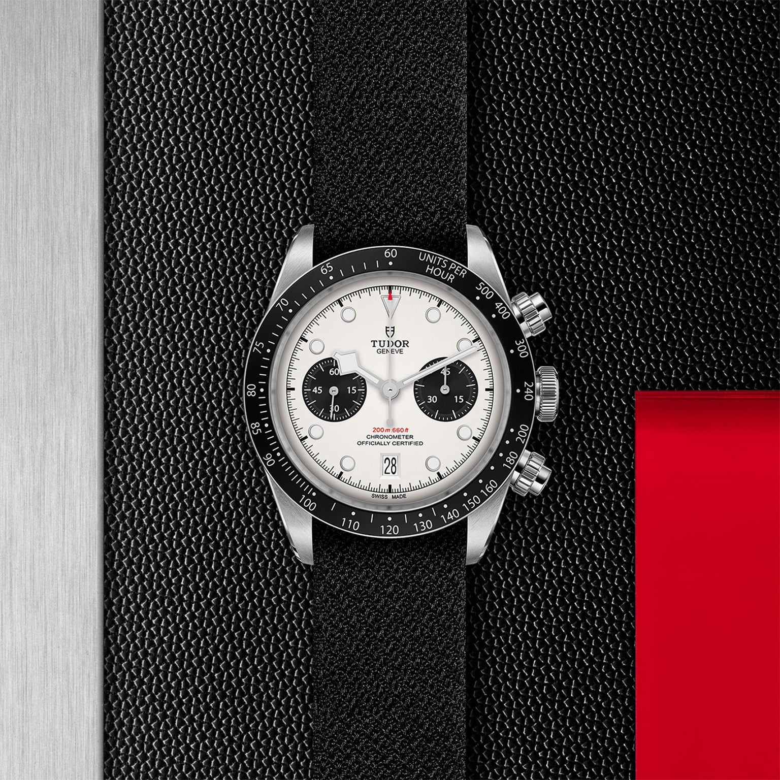 A TUDOR Black Bay Chrono wristwatch with a white dial and black tachymeter bezel sits on a textured black surface next to a metallic panel and red accent.
