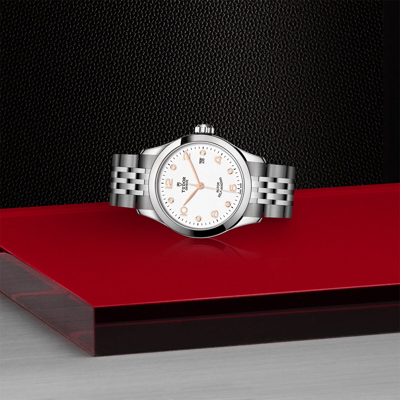 A TUDOR 1926 silver wristwatch with a white dial and metal bracelet is showcased on a red and black platform against a textured black background.