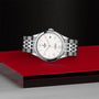 A TUDOR 1926 wristwatch with a silver metal bracelet and white dial is showcased on a glossy red and black surface, set against a textured black background.