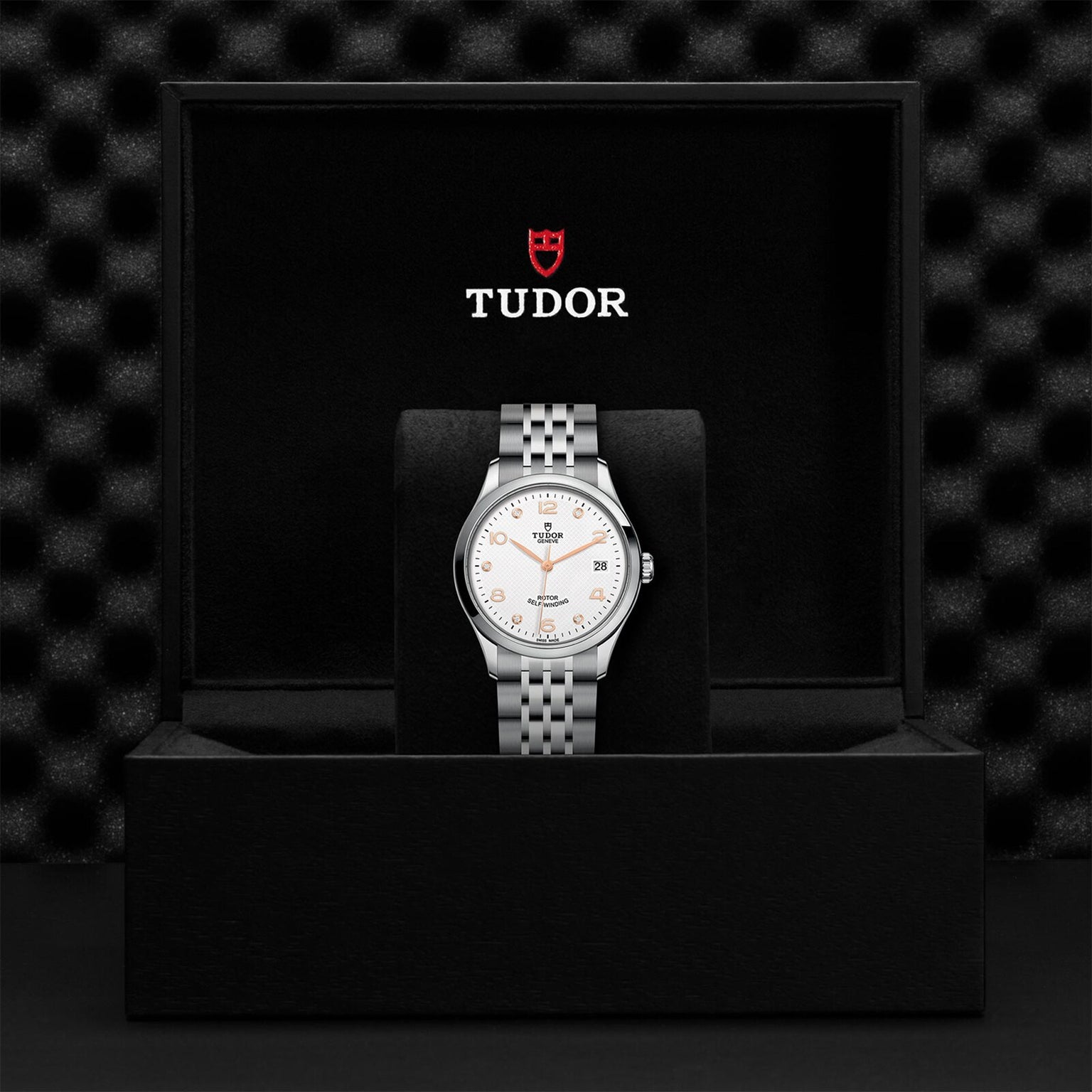 A TUDOR 1926 silver wristwatch with a white dial is displayed inside an open black TUDOR presentation box.