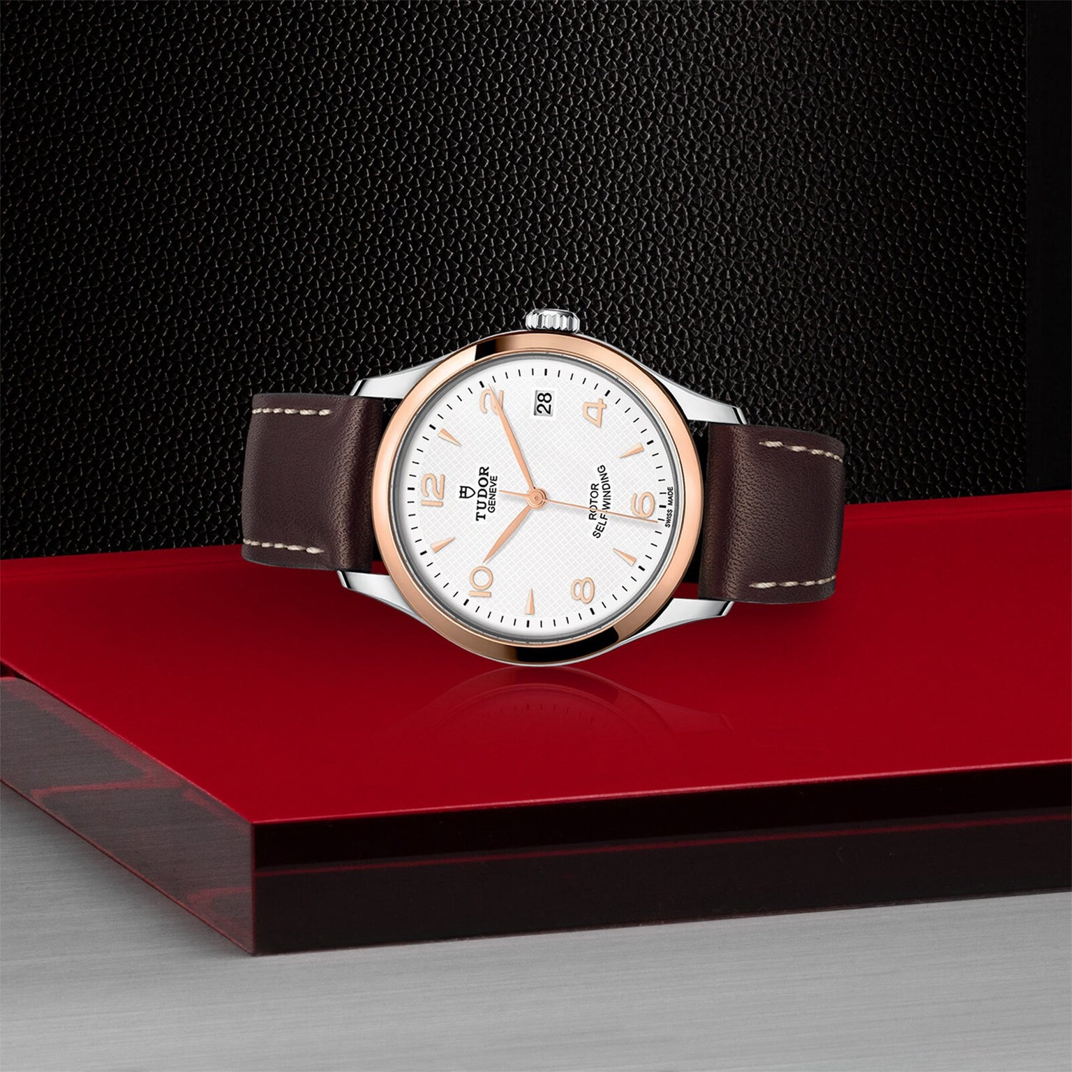 A TUDOR 1926 wristwatch with a brown leather strap and white dial, displayed on a red and black platform.