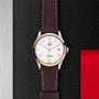 The TUDOR 1926 wristwatch features a white dial, brown leather strap, and silver case, elegantly displayed on a textured black surface with metallic and red accents.