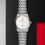 The TUDOR 1926 is a timeless silver wristwatch featuring a white dial, gold hour markers, date display, and metal bracelet, shown on a black and steel surface with a red accent. Perfect for lovers of mechanical watches.