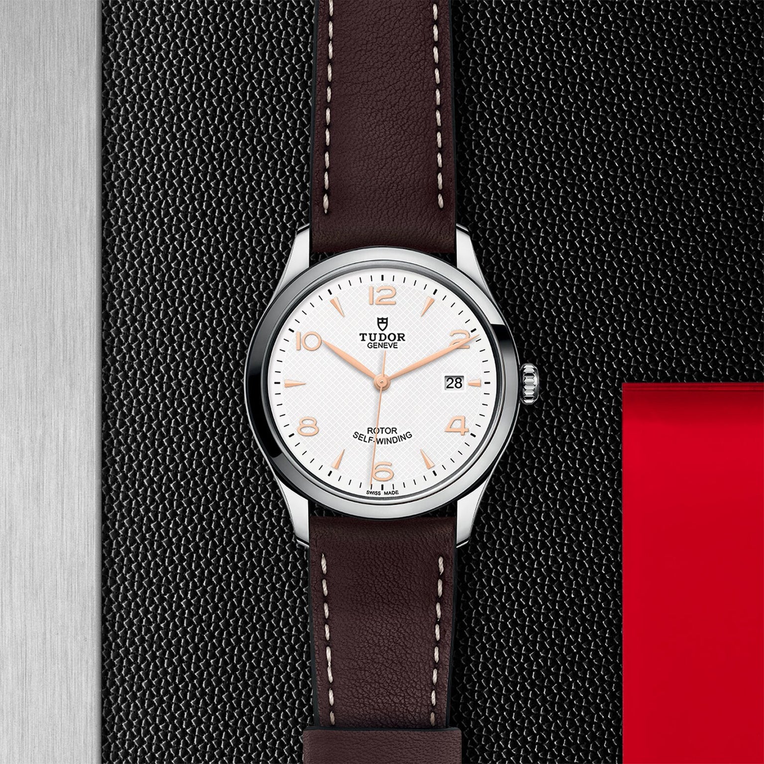 The TUDOR 1926 wristwatch, featuring a white dial, brown leather strap, silver case, and date display, rests on a black textured surface with a red and metallic backdrop—highlighting the classic allure of mechanical timepieces.