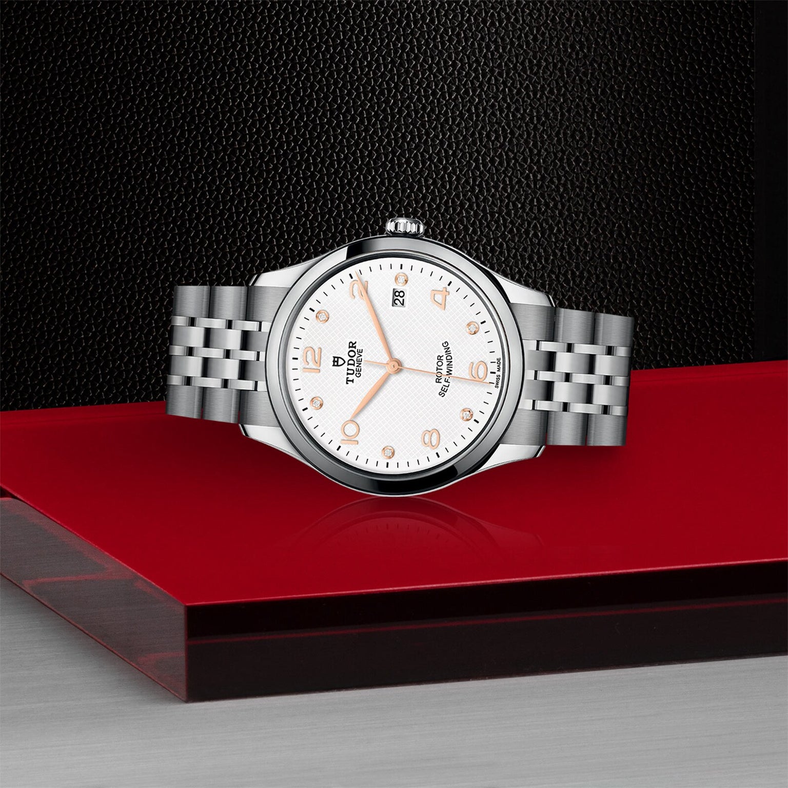 The TUDOR 1926, a silver wristwatch with a white dial, gold hour markers, and date display—symbolizing classic elegance—is displayed on a striking red and black geometric platform.