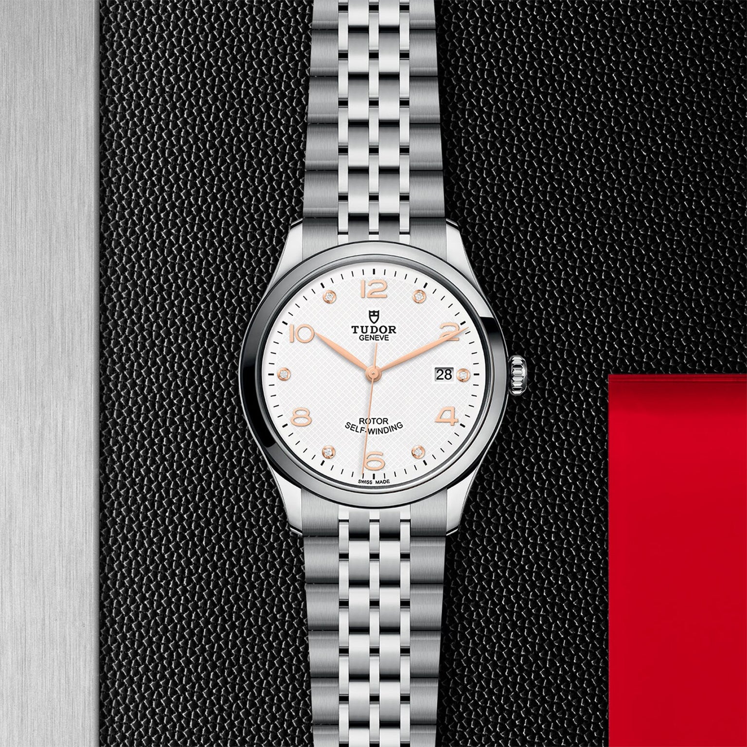 The TUDOR 1926 is a stainless steel wristwatch with a white dial, gold hour markers, and date display, elegantly displayed on a textured black and red background—showcasing classic Tudor style.