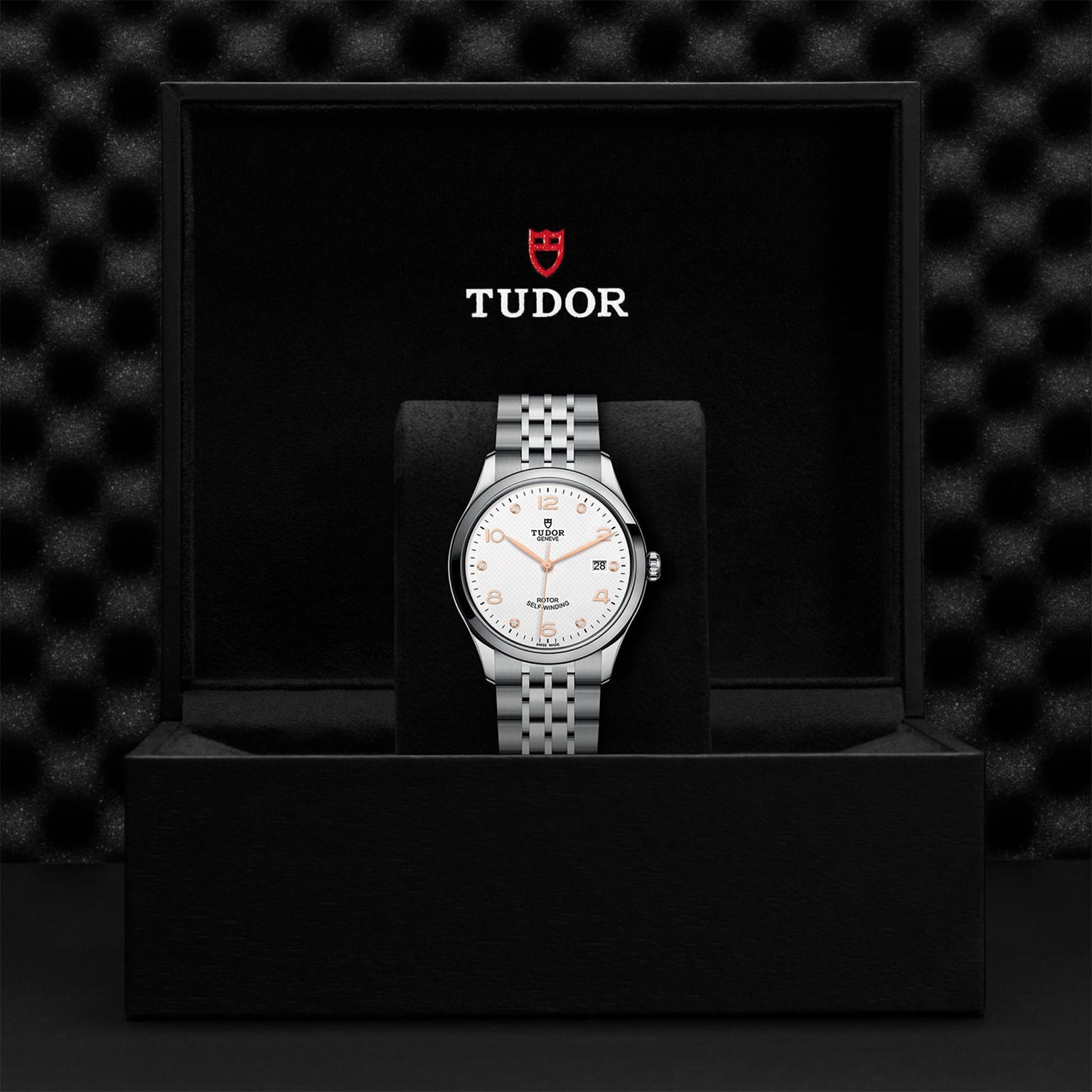 The TUDOR 1926 wristwatch, featuring a silver metal band and white dial, is showcased upright in an open black box with TUDOR branding.