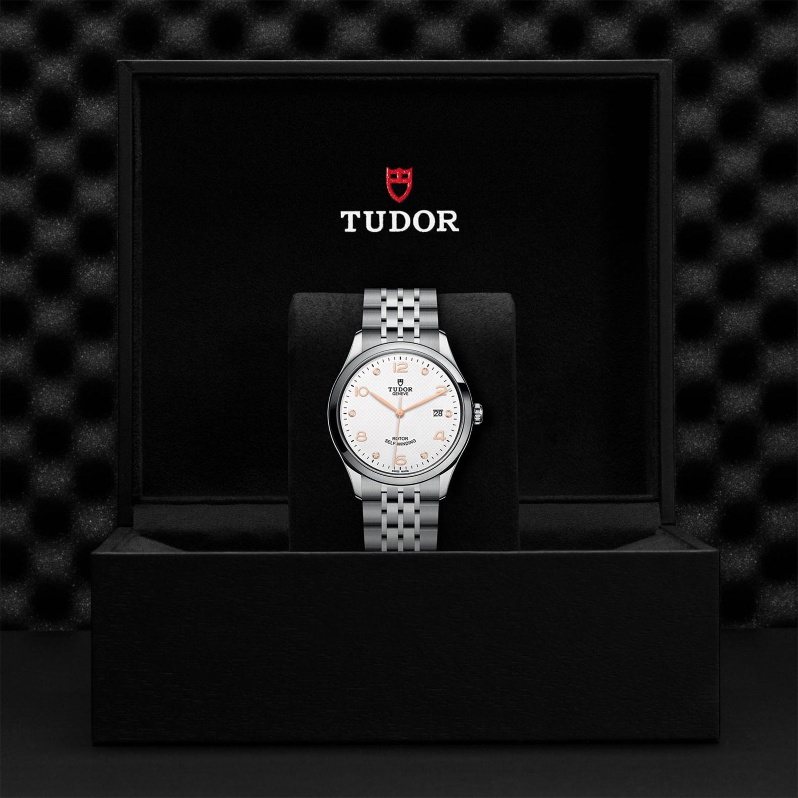 The TUDOR 1926 wristwatch, featuring a silver metal band and white dial, is showcased upright in an open black box with TUDOR branding.