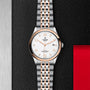 A TUDOR 1926 wristwatch with a white dial, gold bezel, date display, and a gold & silver metal bracelet rests on a textured black and red surface.