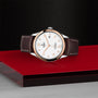 The TUDOR 1926 wristwatch features a white dial, gold hour markers, a brown leather strap, and date display—exemplifying classic elegance. It’s presented on a red and black platform against a textured black background.