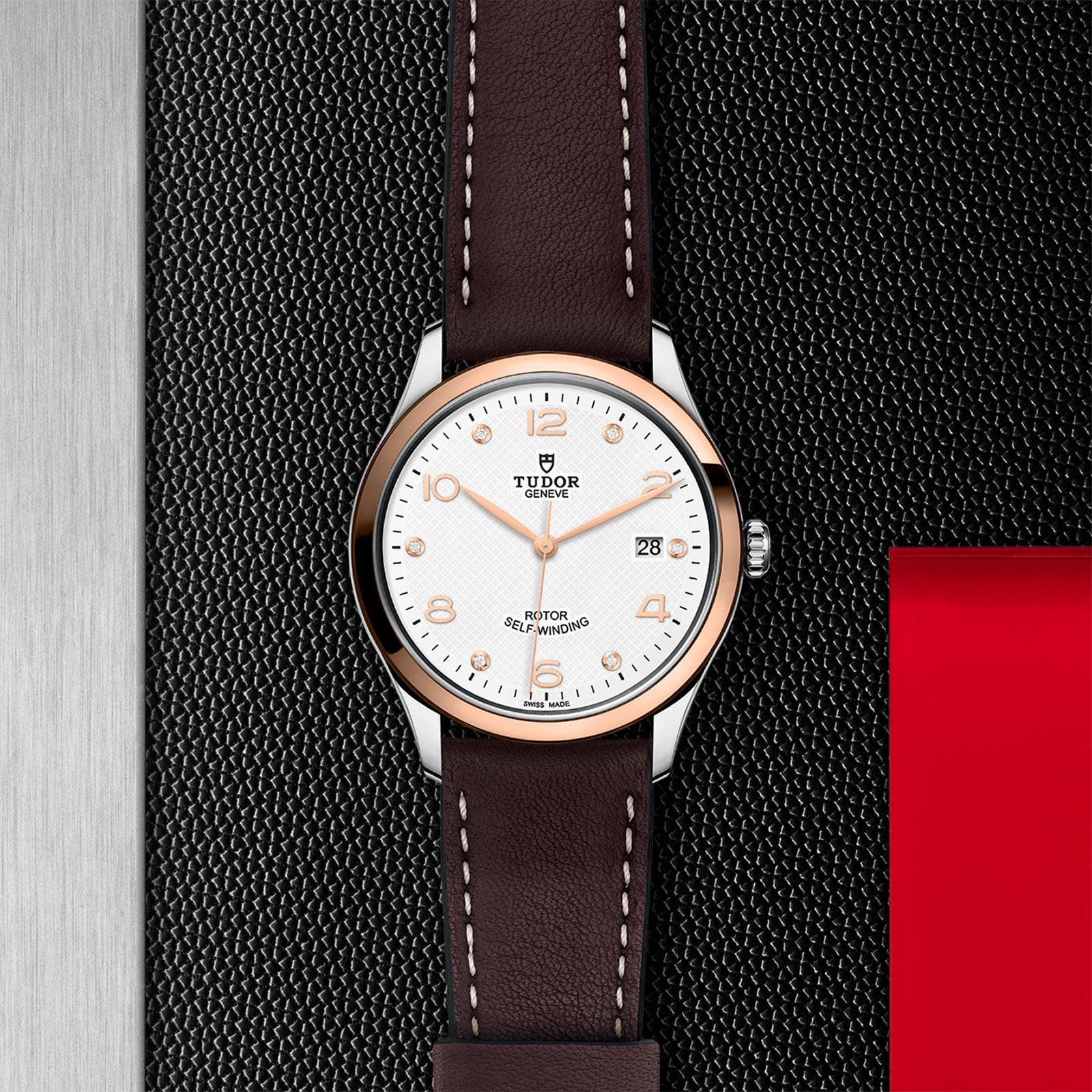 The TUDOR 1926 wristwatch, featuring a white dial, gold indices, date display, and brown leather strap, is displayed on a textured black and metallic surface.