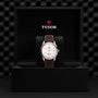 The TUDOR 1926 wristwatch, with a white dial and brown leather strap, is displayed upright in an open black box featuring the TUDOR logo.