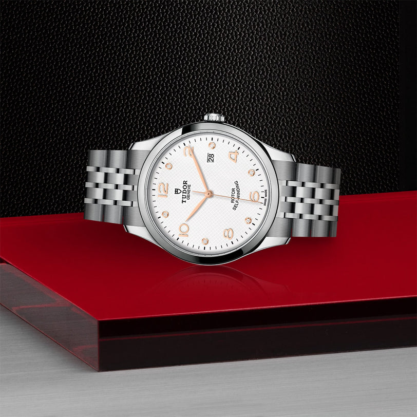 A TUDOR 1926 wristwatch with a silver case, white dial, gold markers, and a metal bracelet is displayed on a red and black surface against a textured dark background, showcasing classic elegance.