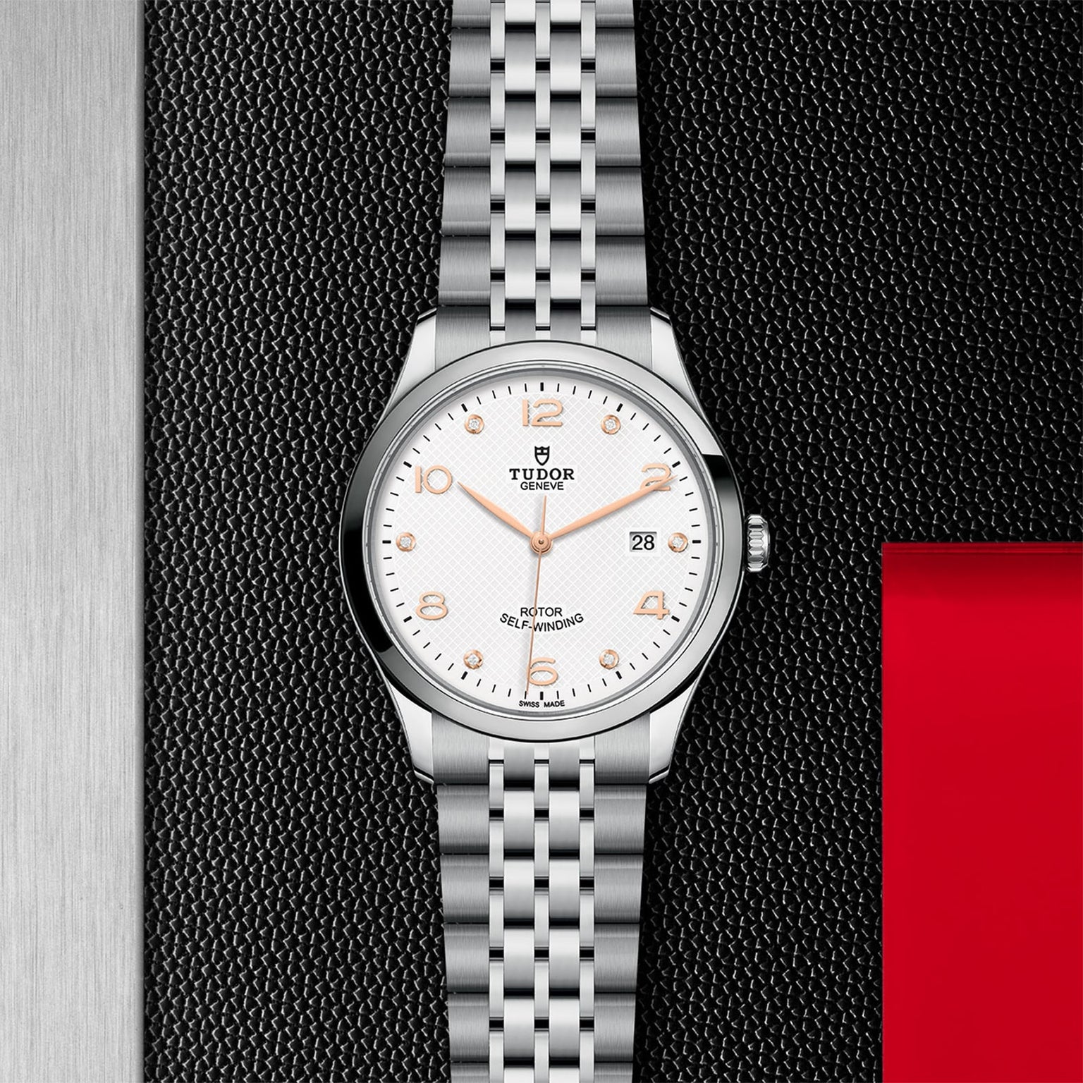 A silver TUDOR 1926 wristwatch with a white dial, gold hour markers, and a metal bracelet is shown on a textured black and red background, accentuating its classic and elegant design.