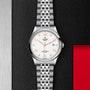 A silver TUDOR 1926 wristwatch with a white dial, gold hour markers, and a metal bracelet is shown on a textured black and red background, accentuating its classic and elegant design.