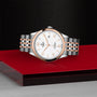 A TUDOR 1926 wristwatch with a silver and rose gold metal band, white dial, and date display rests elegantly on a glossy red and black surface—a refined homage to classic mechanical timepieces.