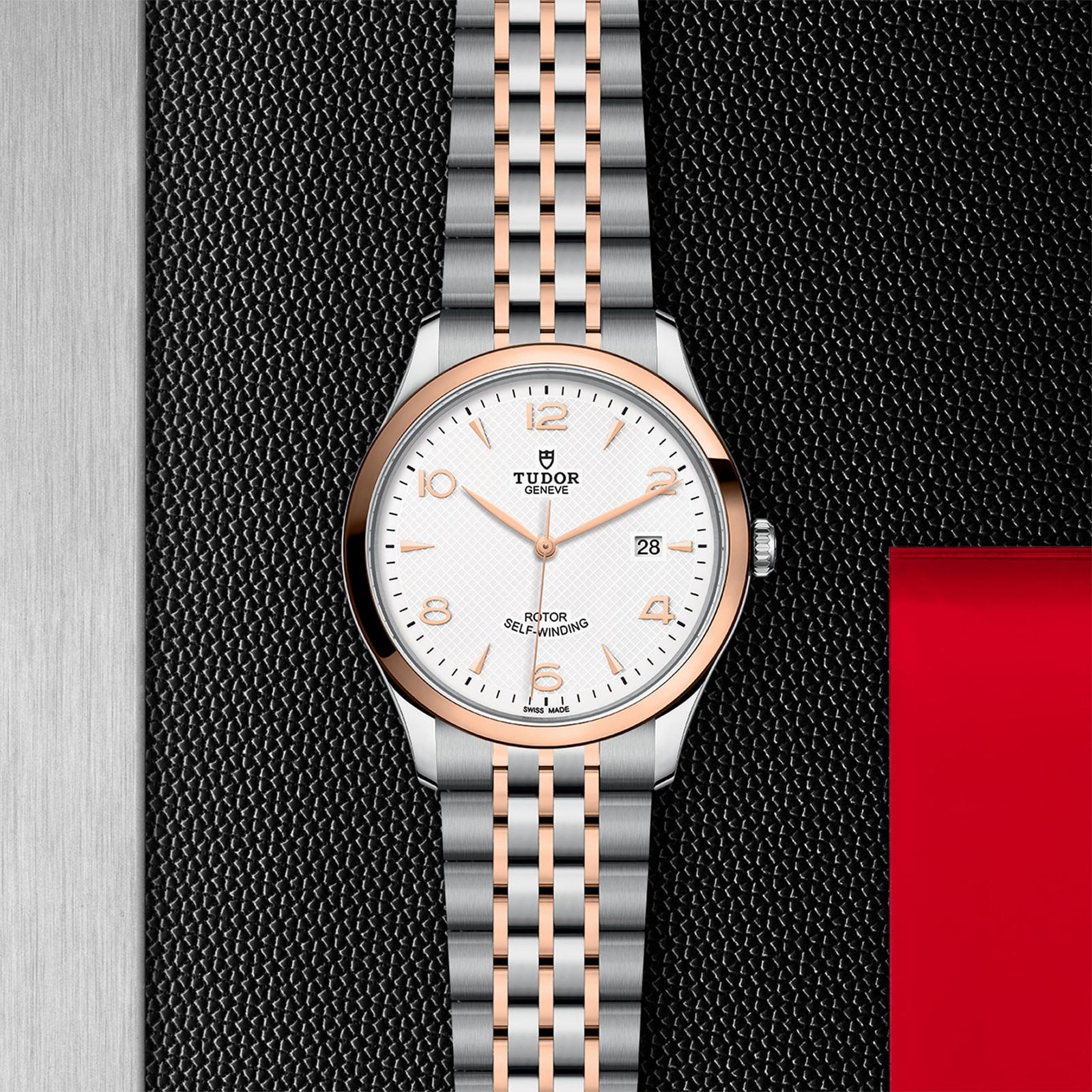 A TUDOR 1926 wristwatch with a white dial, gold accents, and a two-tone bracelet is displayed on a textured black surface bordered in red and silver.