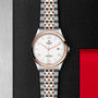 A TUDOR 1926 wristwatch with a white dial, gold accents, and a two-tone bracelet is displayed on a textured black surface bordered in red and silver.