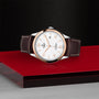 A TUDOR 1926 wristwatch with a white dial, gold accents, and brown leather strap is showcased on a red and black platform against a textured dark background, embodying the spirit of mechanical watches.