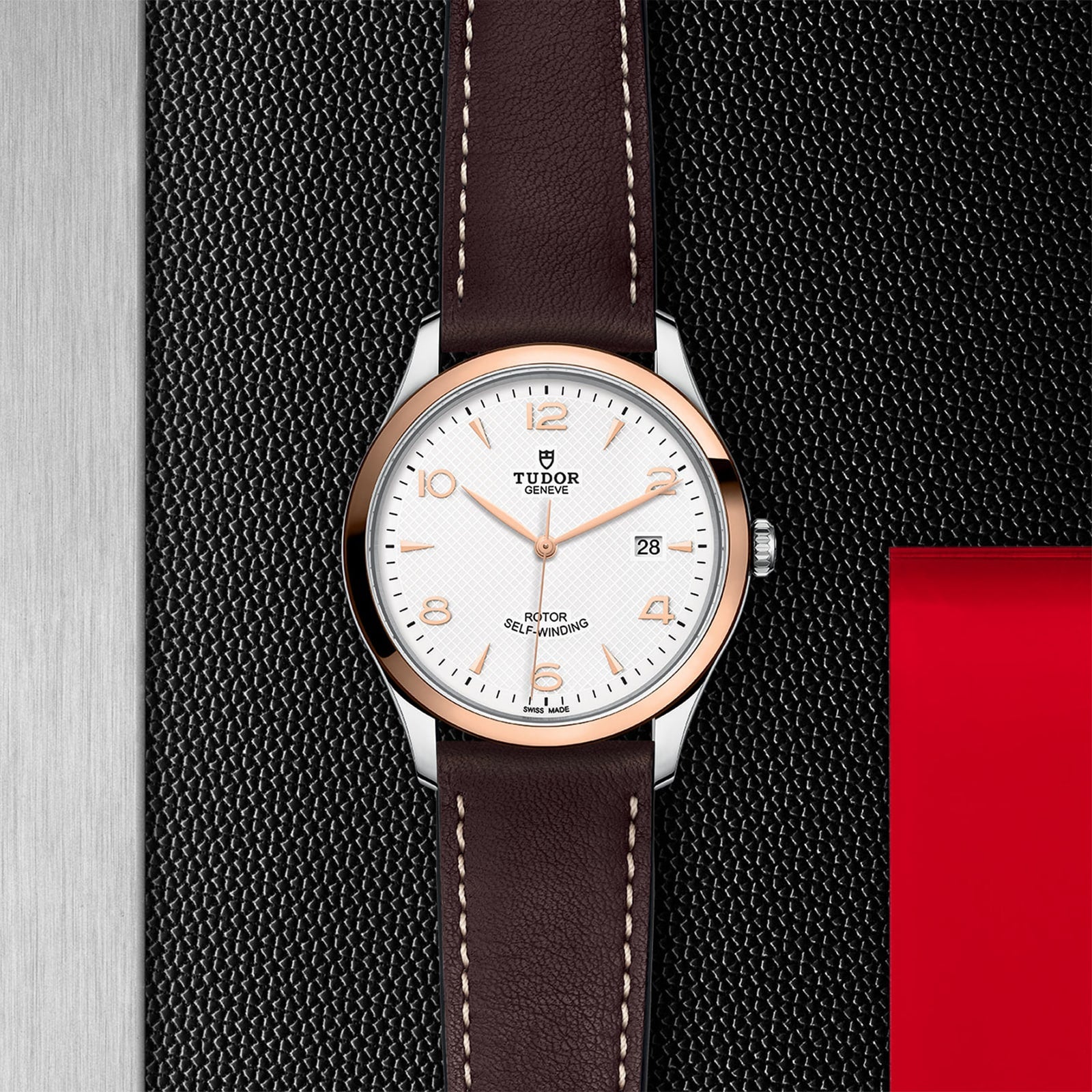 A TUDOR 1926 wristwatch with a white dial and brown leather strap displayed on a textured black and silver background.