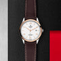 A TUDOR 1926 wristwatch with a white dial and brown leather strap displayed on a textured black and silver background.