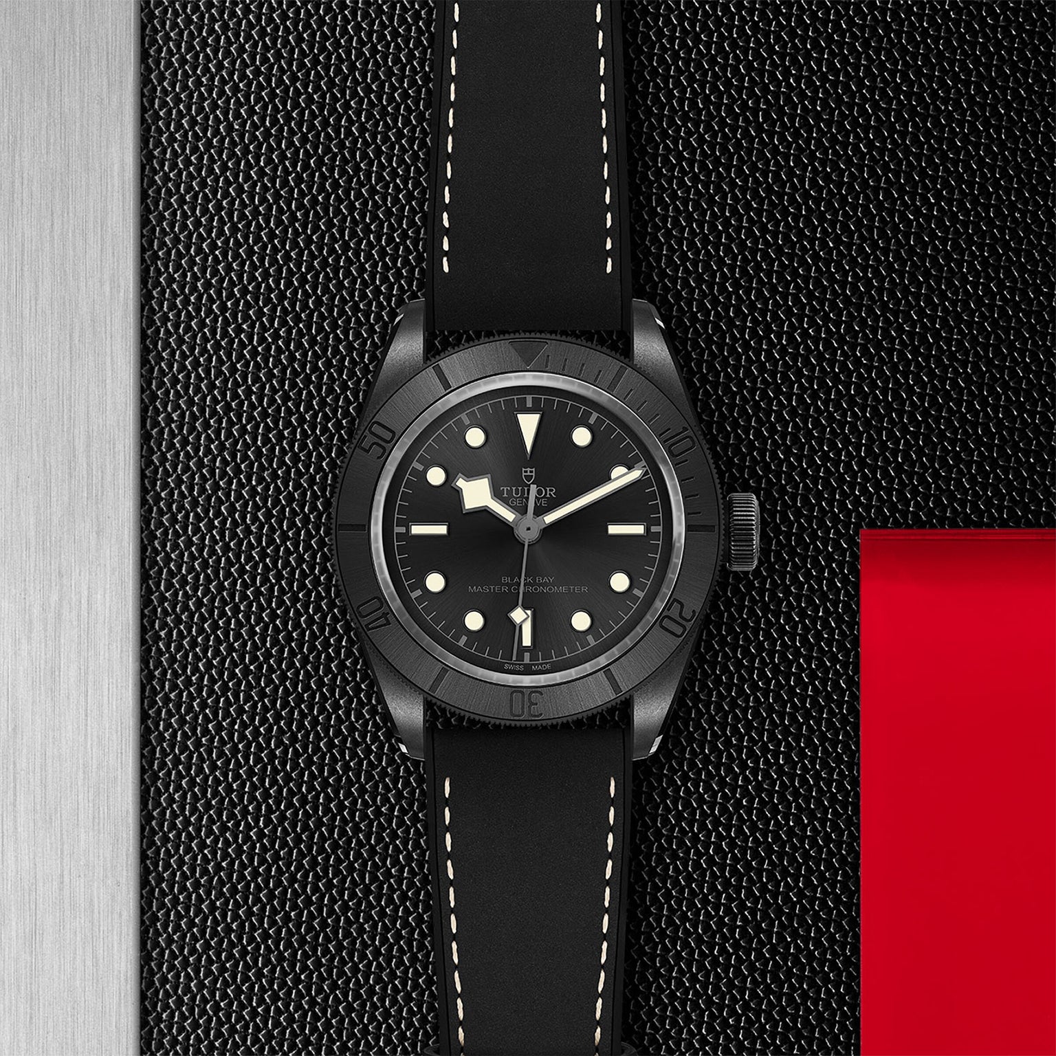 TUDOR Black Bay Ceramic wristwatch with a black strap, displayed on a textured black and red background.