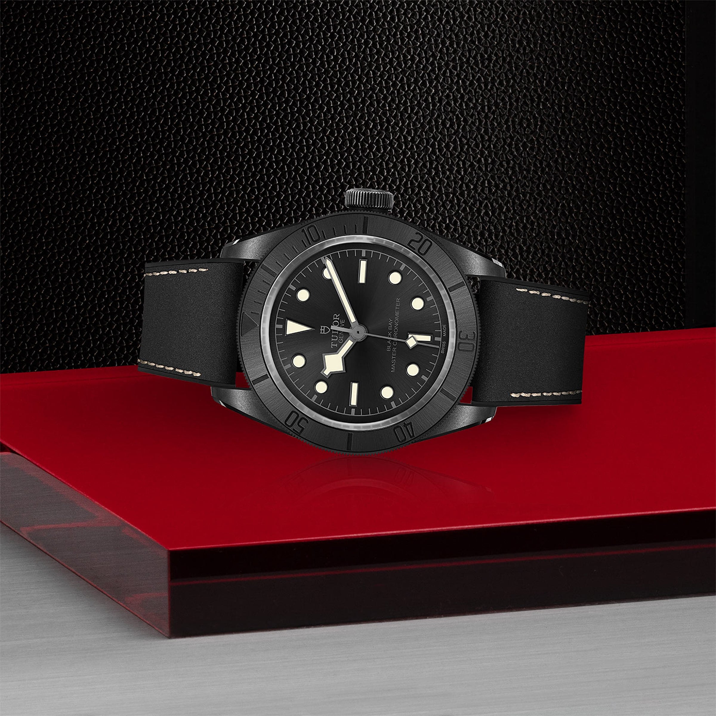The TUDOR Black Bay Ceramic wristwatch, featuring a black face with white hour markers, is showcased on a sleek black and red platform, highlighting the Swiss-made watchs timeless elegance against a textured backdrop.