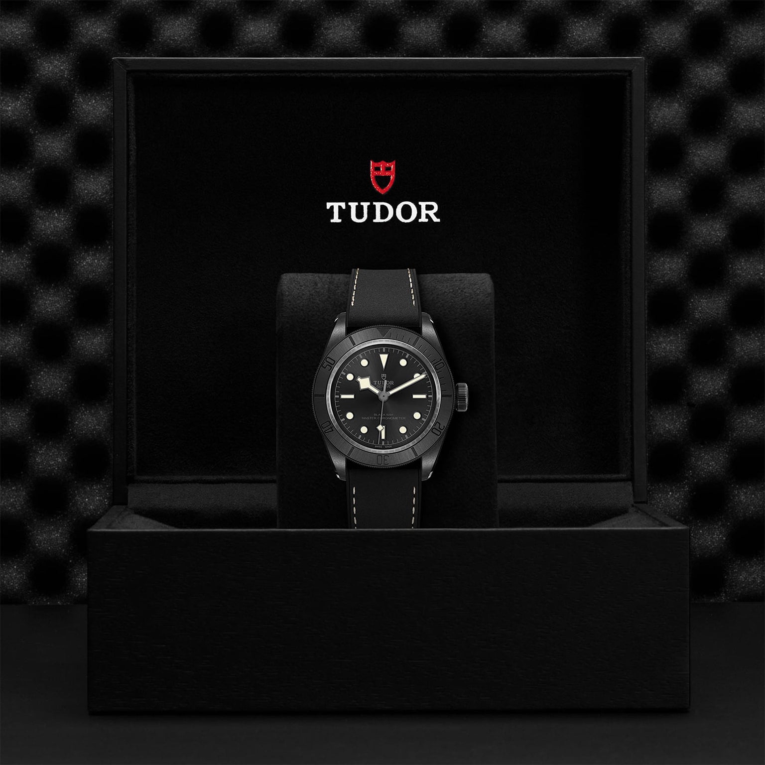 A Swiss-made TUDOR Black Bay Ceramic wristwatch with a black strap is displayed in an open black box, featuring the TUDOR logo and branding on the inside of the lid.