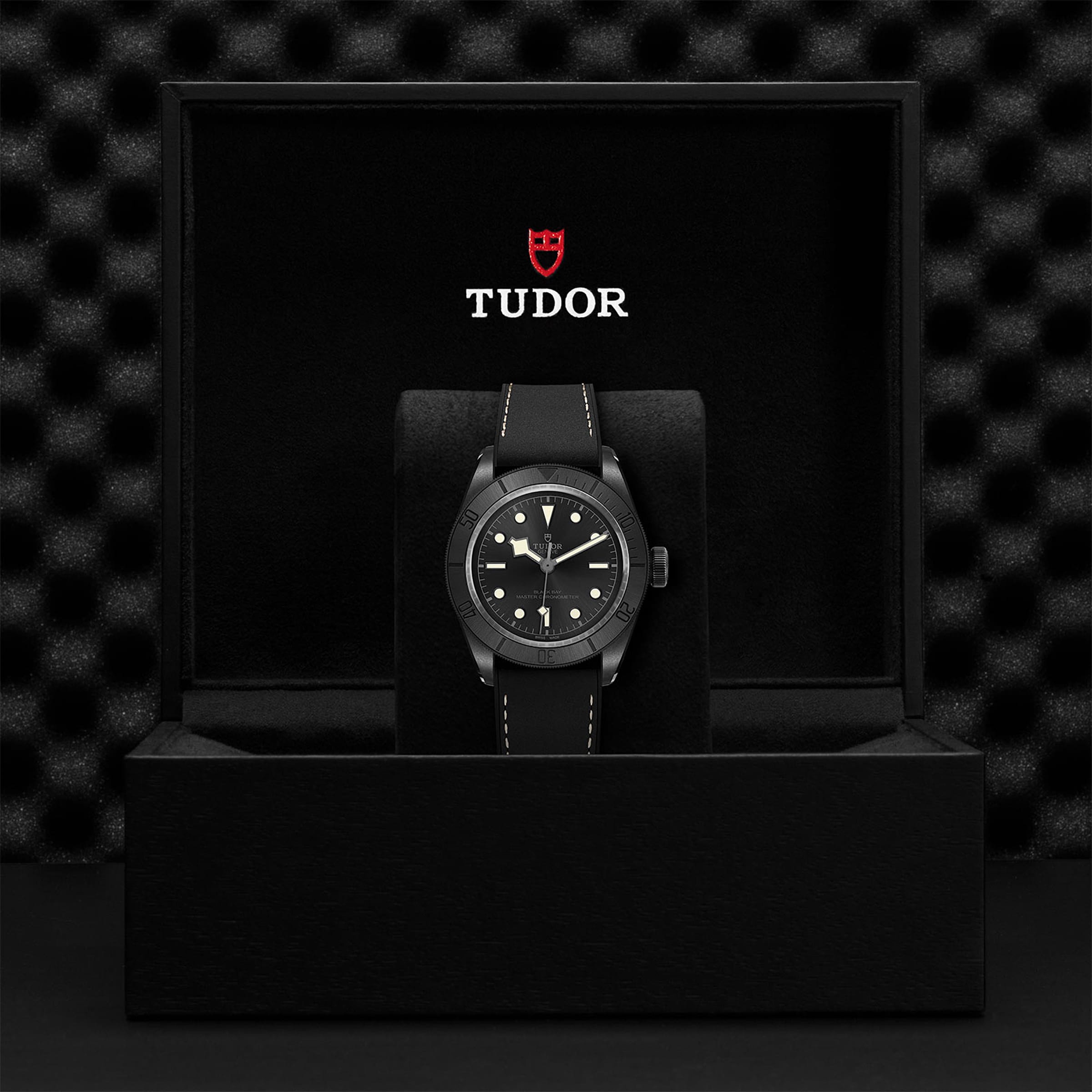 A Swiss-made TUDOR Black Bay Ceramic wristwatch with a black strap is displayed in an open black box, featuring the TUDOR logo and branding on the inside of the lid.