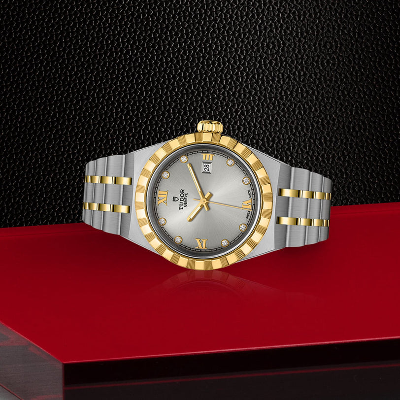 A TUDOR Royal luxury two-tone gold and silver wristwatch displayed on a red and black textured surface.