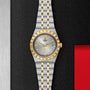 A TUDOR Royal wristwatch with a silver dial, gold hour markers, fluted bezel, and two-tone bracelet rests on a textured black surface.