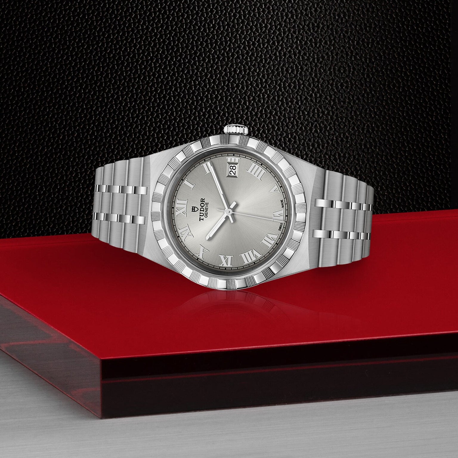 A TUDOR Royal wristwatch with a metal band and Roman numeral markers is displayed on a red and gray platform against a black textured background.