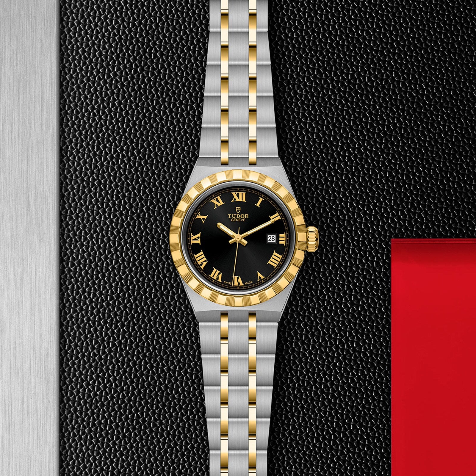A TUDOR Royal wristwatch in stainless steel and gold, featuring a black dial with gold Roman numerals, is displayed on a textured black surface accented with metal and red.