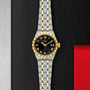 A TUDOR Royal wristwatch in stainless steel and gold, featuring a black dial with gold Roman numerals, is displayed on a textured black surface accented with metal and red.