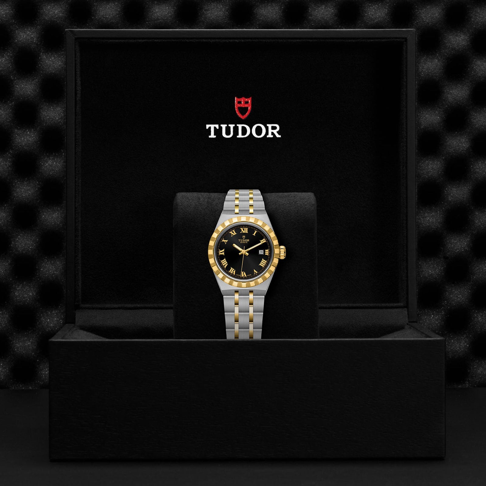 A TUDOR Royal wristwatch, featuring a stainless steel and gold two-tone band with a black-and-gold bezel, is presented in an open Tudor-branded black box, exemplifying the refined sport-chic style of automatic watches.
