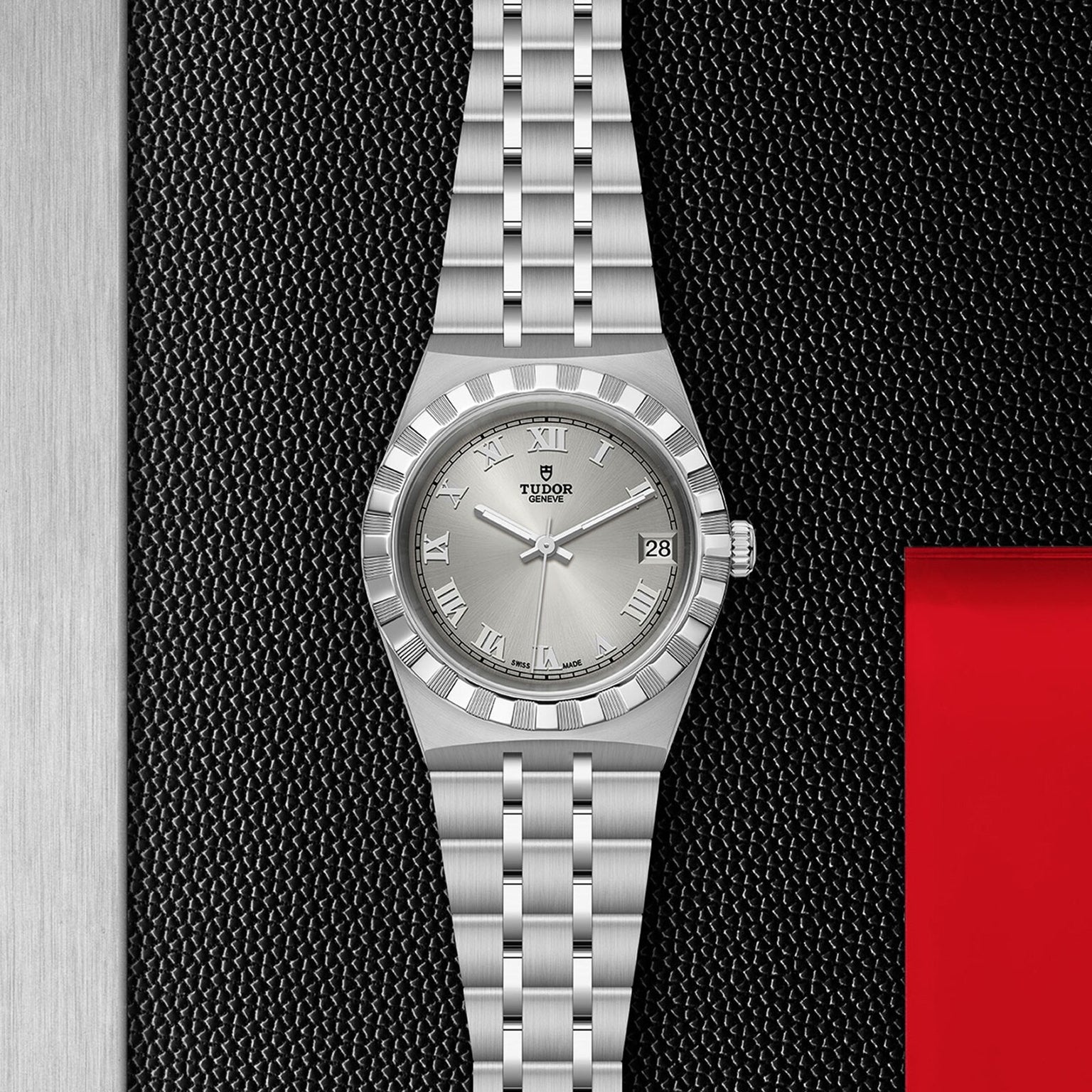 The TUDOR Royal blends stainless steel and gold with Roman numeral markers and a date display, set on a textured black and metallic background with red accents—a striking automatic sport-chic wristwatch.