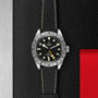 The TUDOR Black Bay Pro features a black dial with luminescent hour markers, date display, and a textured black strap, set against a textured black and silver background with red accents—a modern nod to classic Black Bay design.