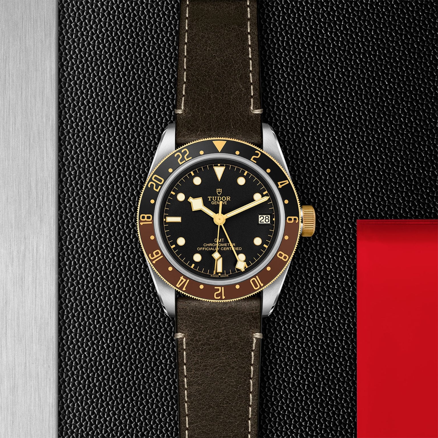 A TUDOR Black Bay GMT S&G watch with a black dial, yellow gold bezel, and black leather strap rests on a textured black and red surface beside a brushed metal panel, highlighting the sophistication of TUDOR’s diver-inspired timepieces.